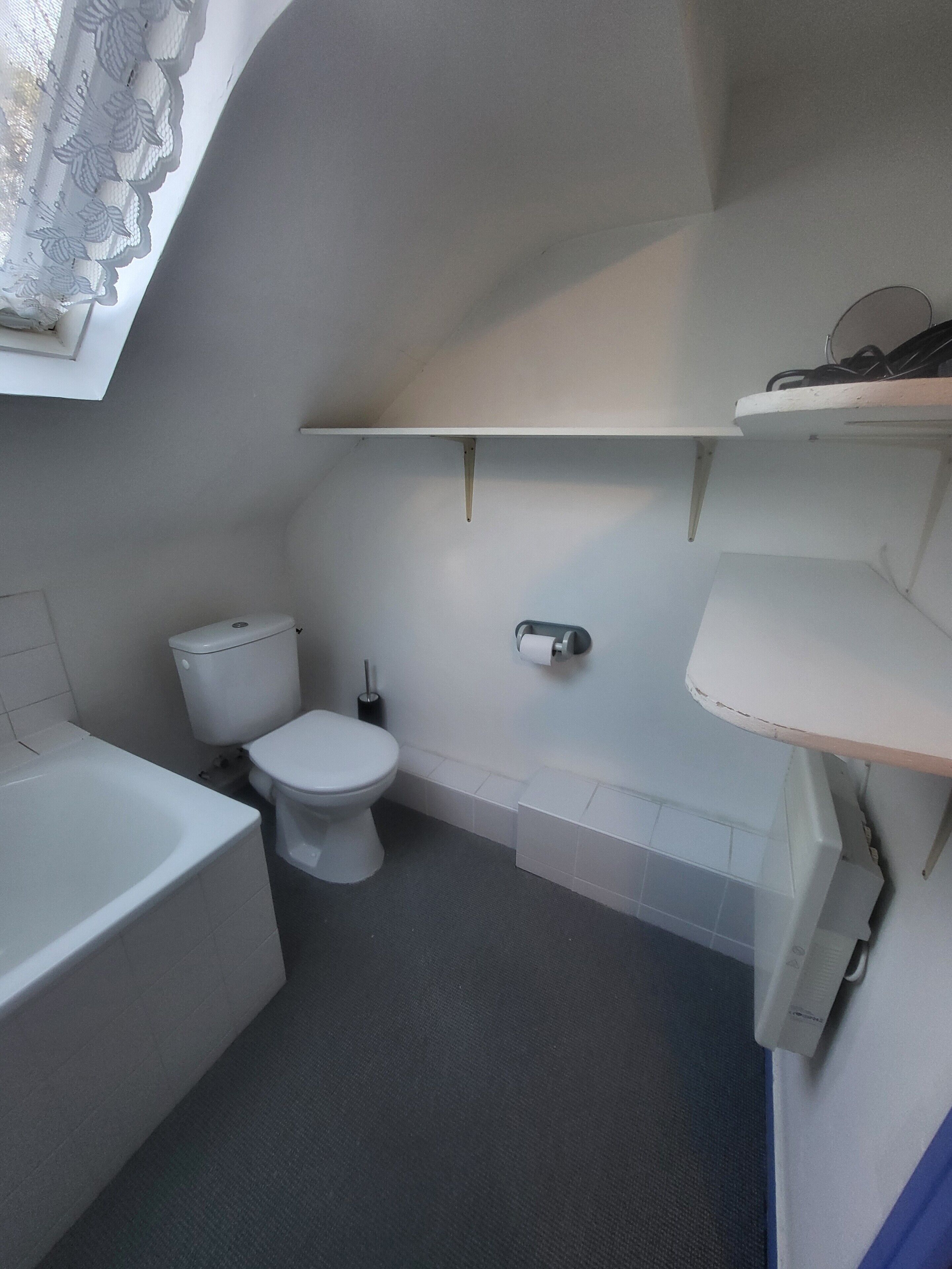 Ein kleines Badezimmer mit Toilette, einer Badewanne und einer Duschkabine. Über der Toilette befindet sich ein Ablagebrett und an der Wand ein Handtuchhalter.