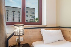 Ein Hotelzimmer mit Bett, Nachttisch, Lampe und Fenster mit Blick auf Gebäude.