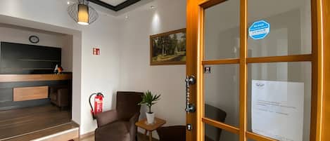 A hotel lobby with a reception desk, seating area, a fire extinguisher, a clock, and a door with a 24-hour service sign.