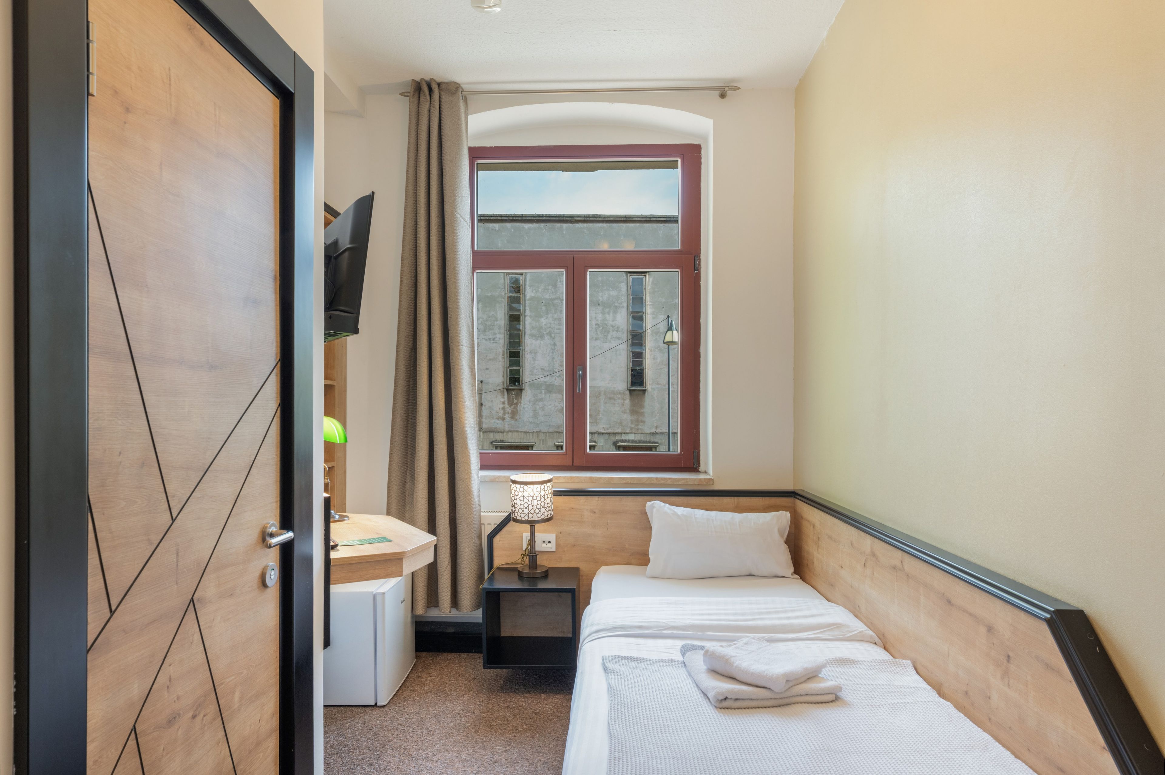 Ein Hotelzimmer mit einem Bett, einem Schreibtisch, einem Fenster mit Vorhängen und einem an der Wand montierten Fernseher.