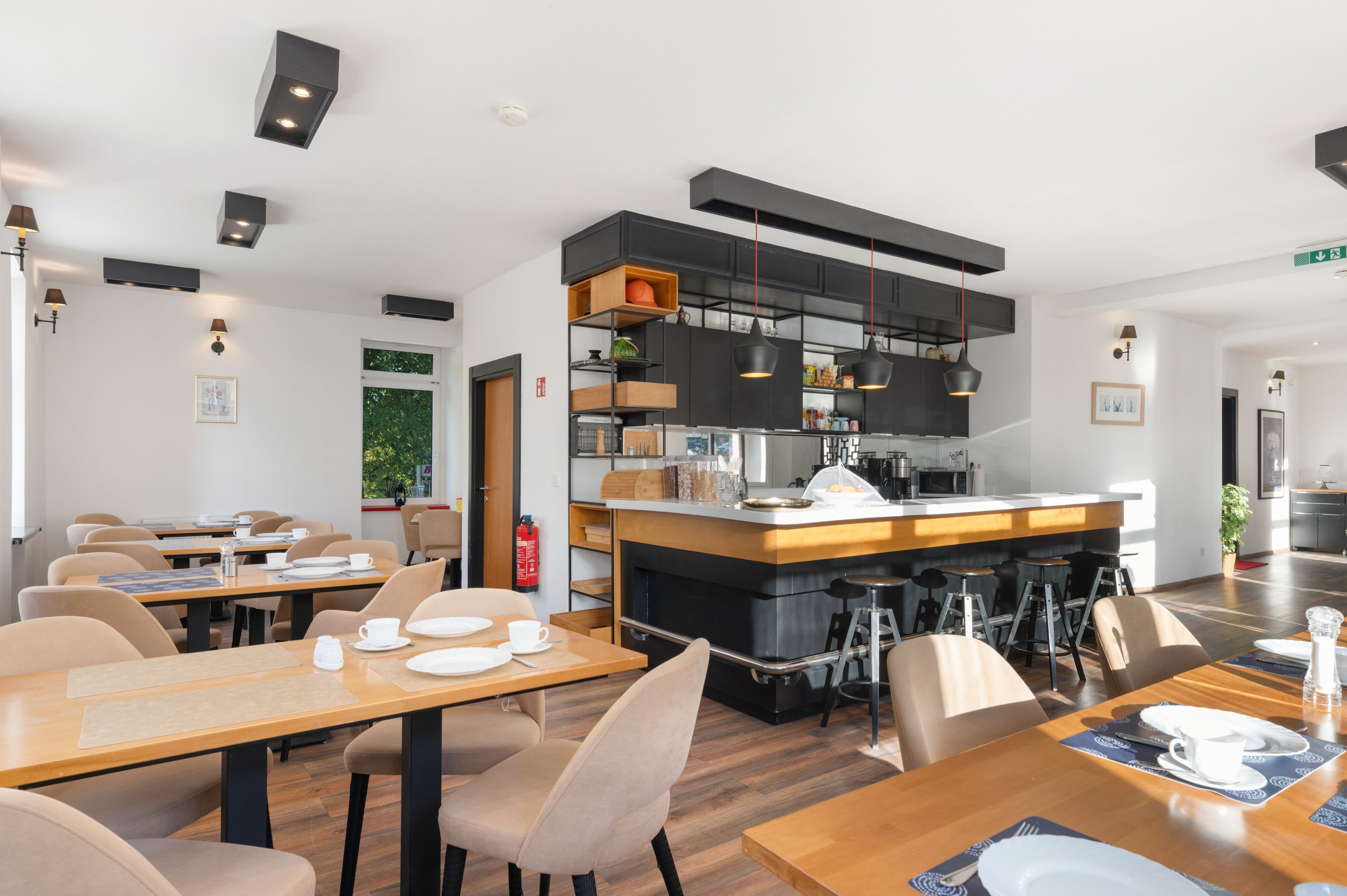 A modern dining area with wooden tables and chairs, a central bar area with stools, and a kitchen with black cabinetry and hanging lights.