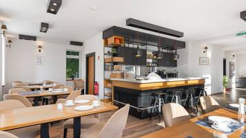 Ein moderner Essbereich mit Holztischen und -stühlen, eine zentrale Bar mit Hockern und eine Küche mit schwarzen Schränken und hängenden Lampen.