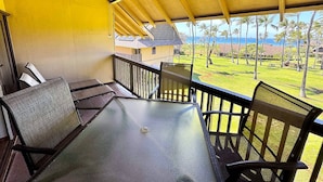A covered outdoor seating area with a table and chairs, overlooking a lawn with palm trees and a view of the ocean.
