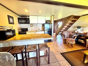 A compact kitchen with a microwave, coffee maker, and a refrigerator. There is a dining area with a small table and chairs. A staircase leads to an upper level.