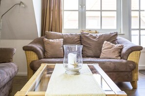 Ein Wohnzimmer mit einem braunen Ledersofa, einer Glasvase mit Kerze und Muscheln auf einem Holztisch sowie einem Fenster mit Vorhängen.