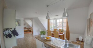 Une cuisine avec un plan de travail en bois, un évier et deux suspensions lumineuses. On y trouve également un coin repas avec des chaises en osier et une table sur laquelle sont disposés des fruits.
