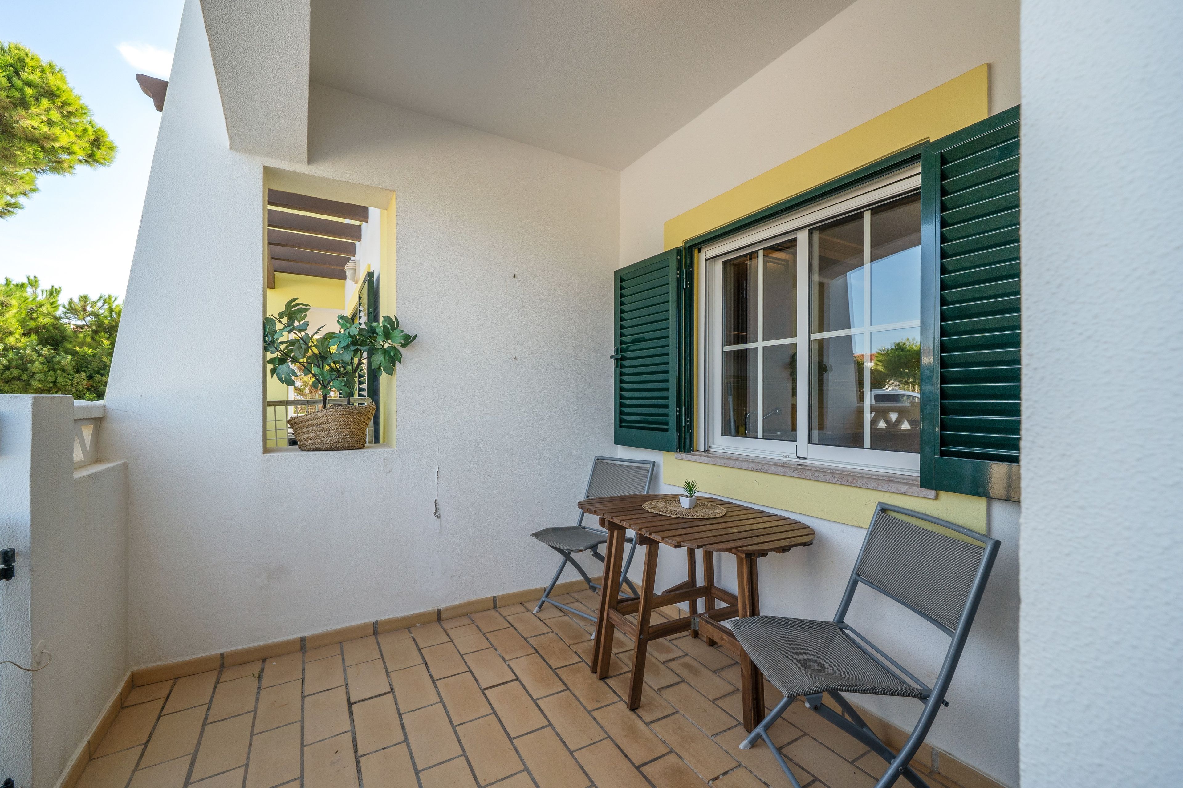 A small patio with a table and two chairs, a window with shutters, and a potted plant.