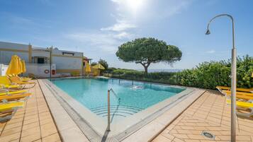 A swimming pool with a tiled deck, yellow sun loungers, and a large tree in the background.