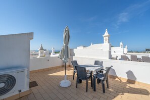 A rooftop terrace with a table and chairs, an air conditioning unit, and a view of the sea and white buildings in the background.
