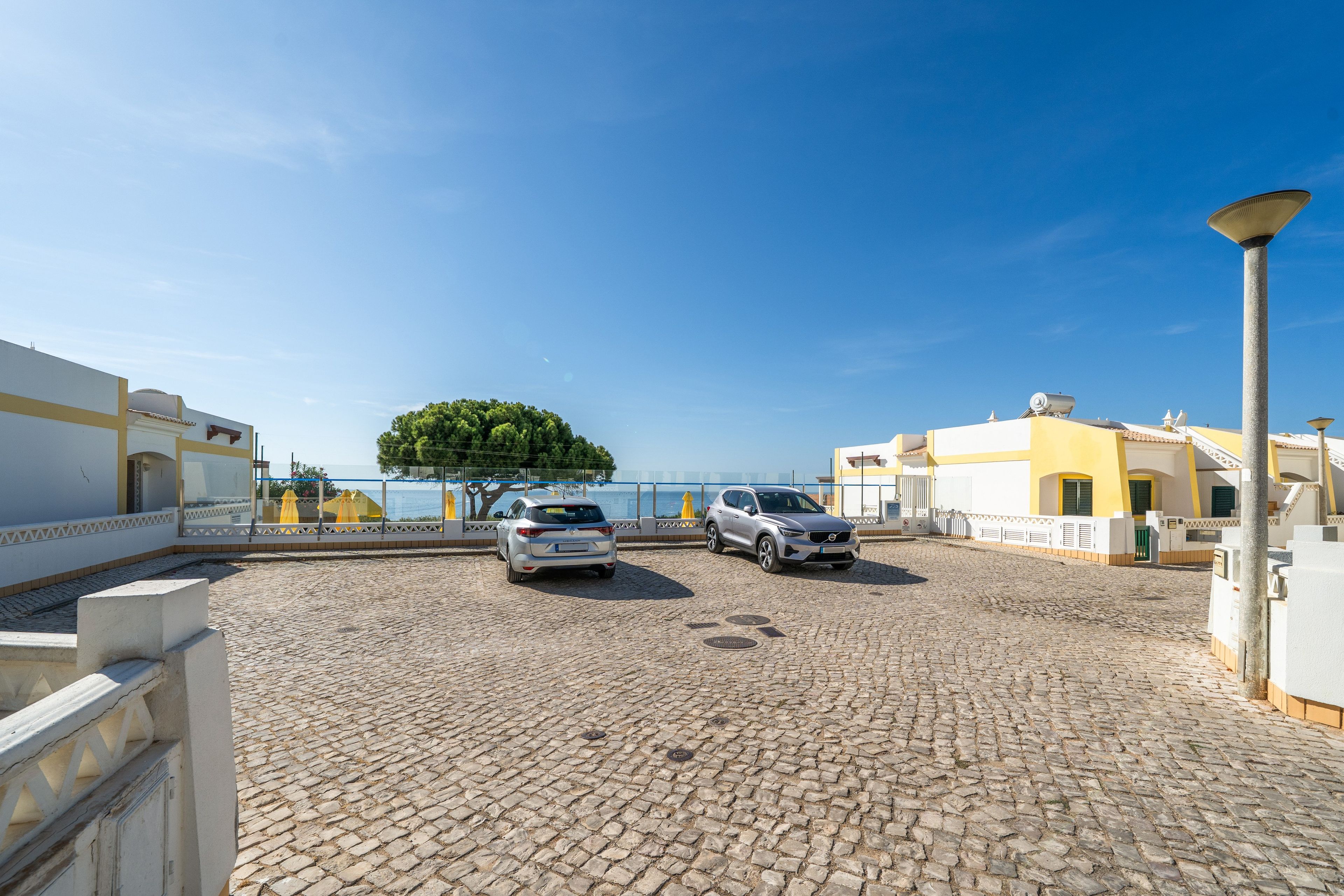 A cobblestone courtyard with two parked cars, a clear blue sky, and a view of the sea.