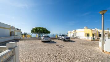 A cobblestone courtyard with two parked cars, a clear blue sky, and a view of the sea.