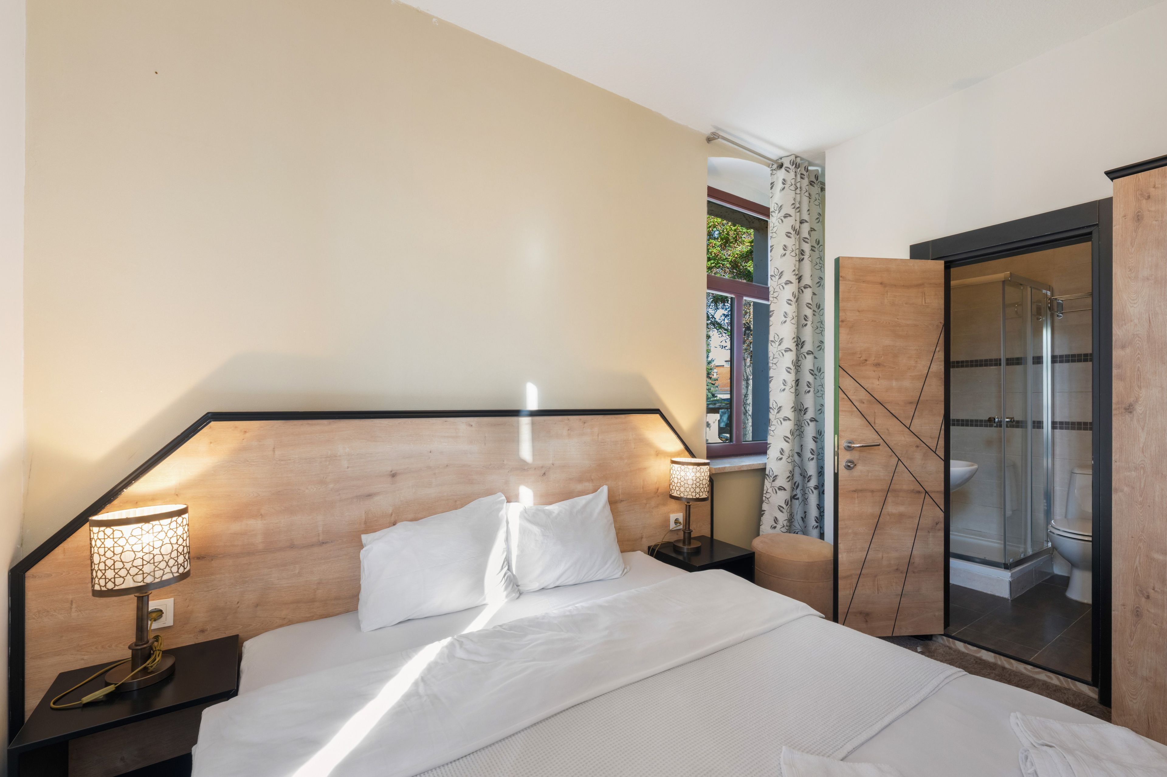 Una habitación de hotel con cama, lámparas de noche, ventana con cortinas y zona de ducha.