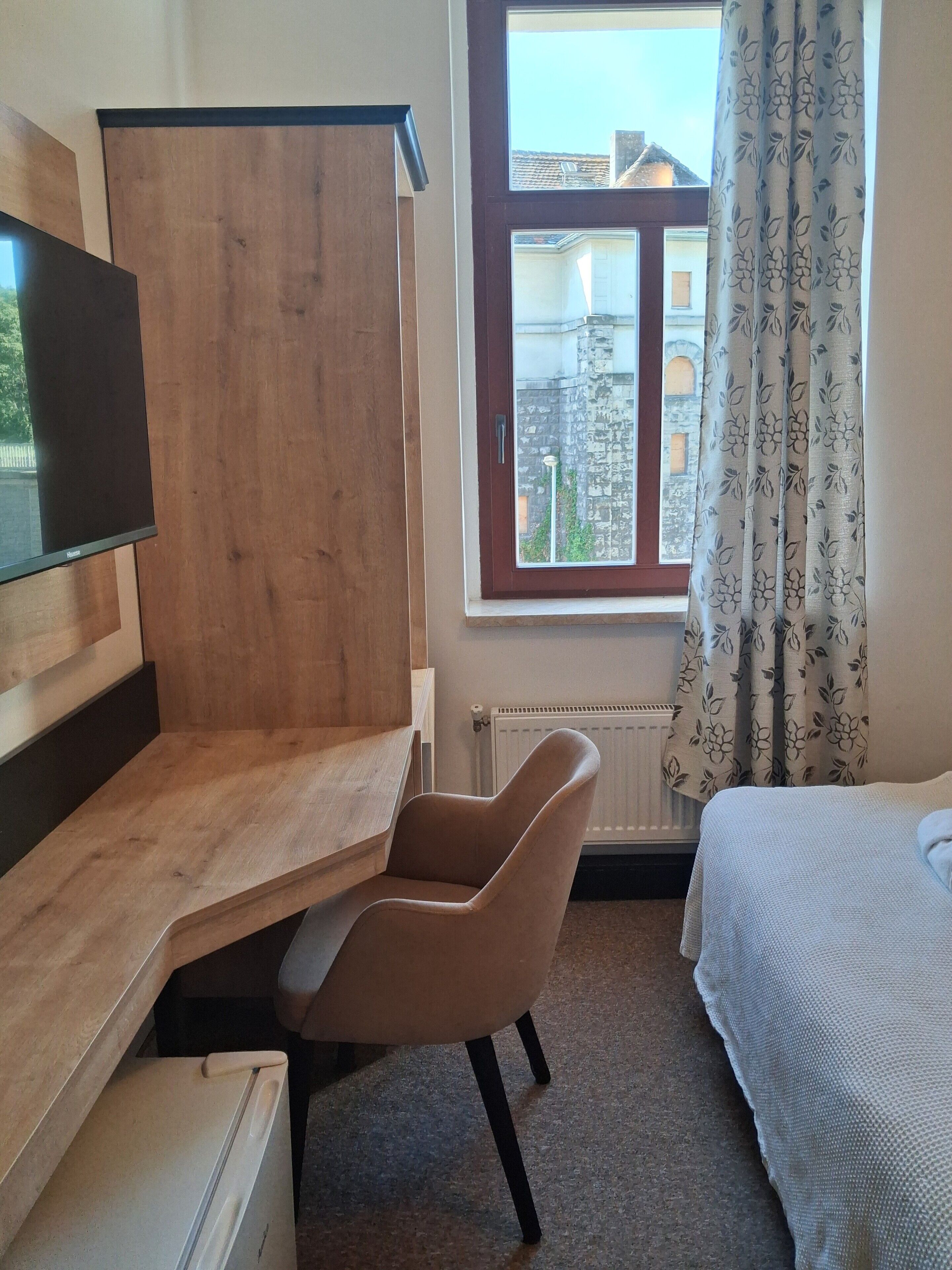 A room with a wooden desk, a chair, a bed, a window with curtains, and a radiator.