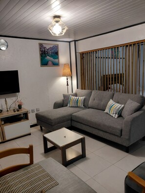 Un salon avec un canapé d’angle gris, une petite table basse blanche, un téléviseur posé sur un meuble en bois, une lampe et un tableau accroché au mur.