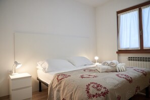 A neatly made bed with a floral-patterned bedspread, a bedside lamp, and rolled towels on the nightstand. - ‘Dolomis Margherita 4px’ Apartment with Mountain View, Shared Garden and Wi-Fi (Caderzone Terme)
