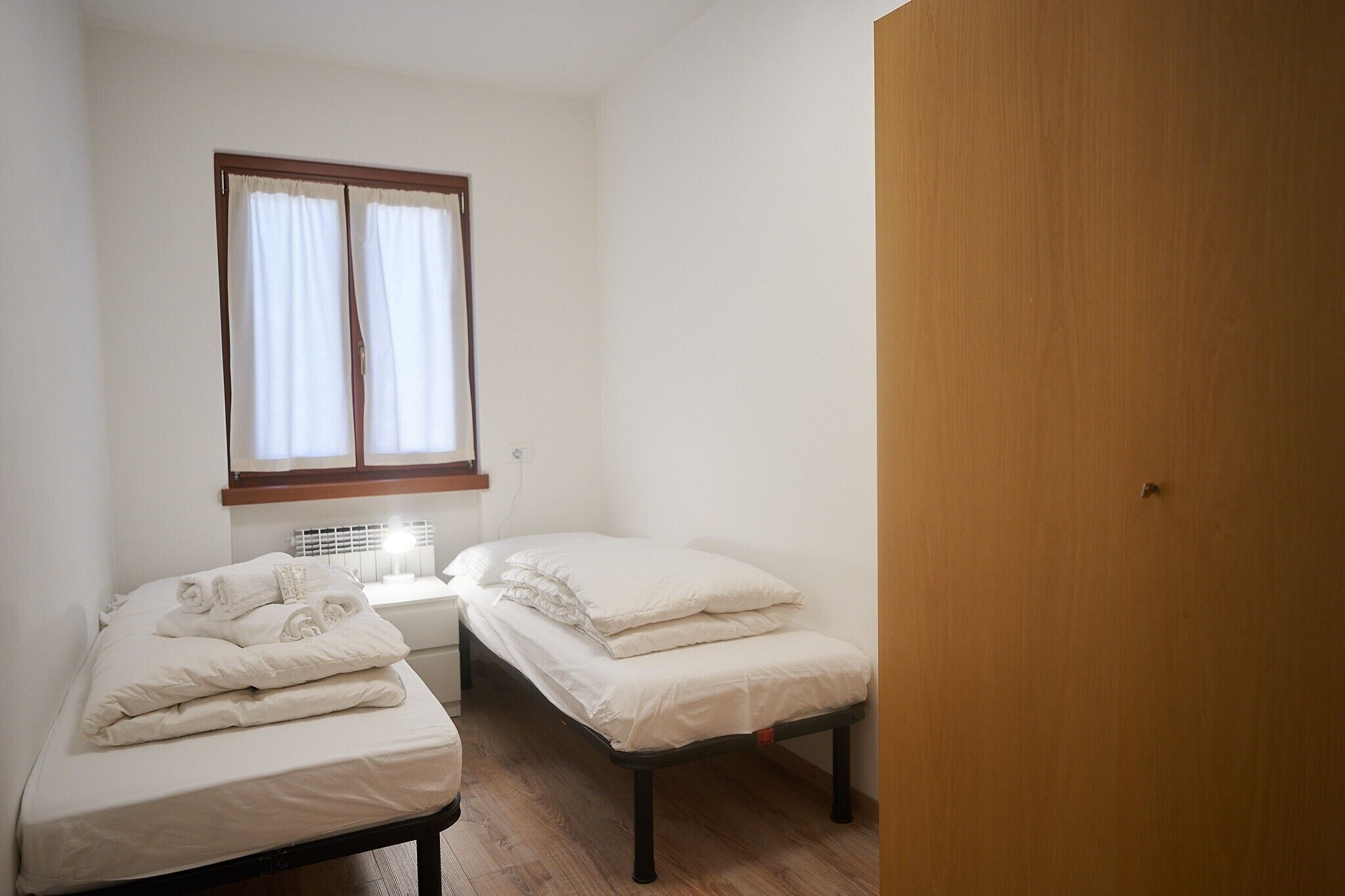 Een kleine, schone kamer met twee bedden, een raam met gordijnen, een nachtkastje met een lamp en een houten deur.