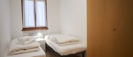 A small, clean room with two beds, a window with curtains, a bedside table with a lamp, and a wooden door.