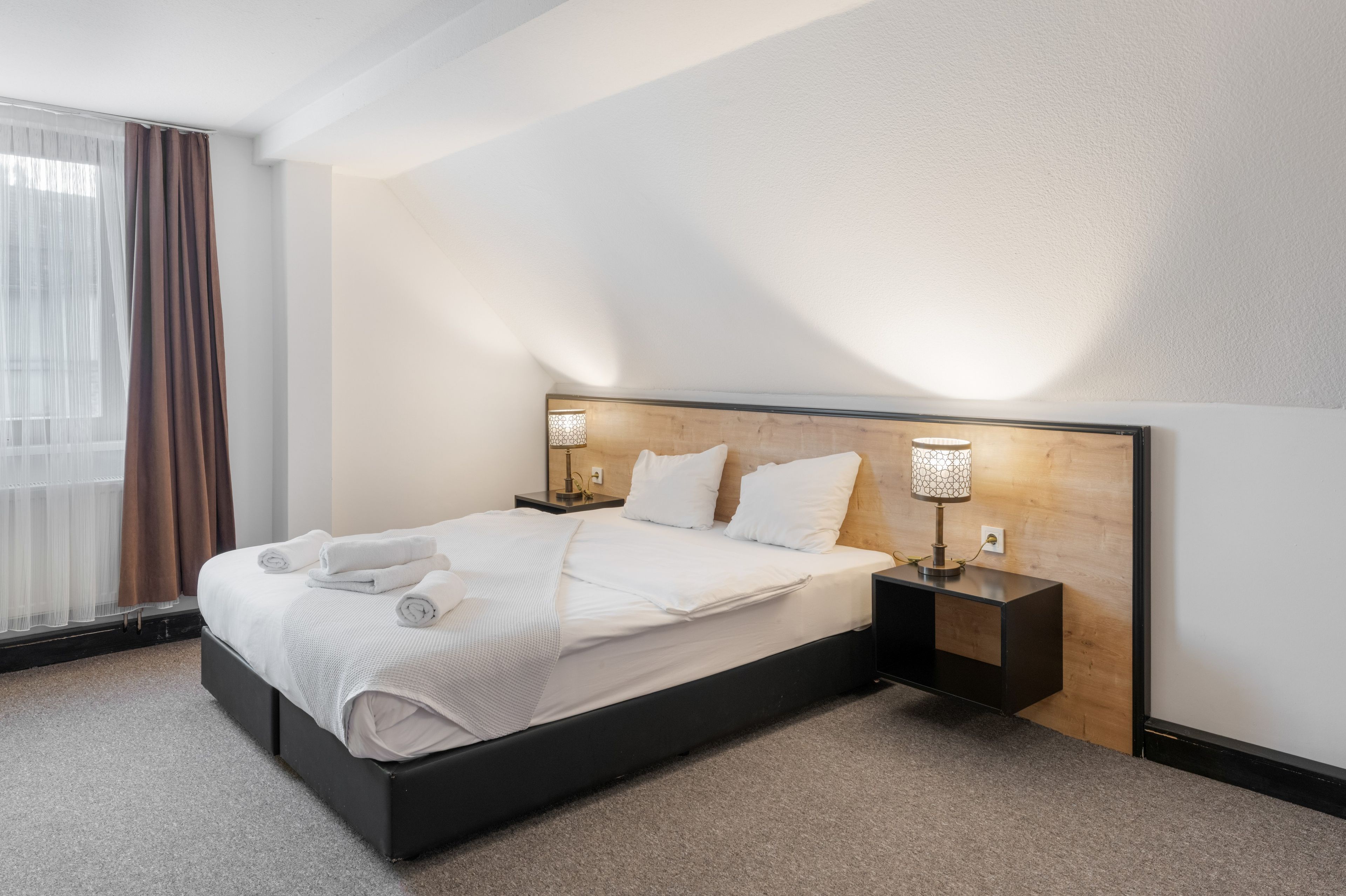 Een hotelkamer met een groot bed, twee nachtkastjes, een raam met gordijnen en een houten hoofdeinde.