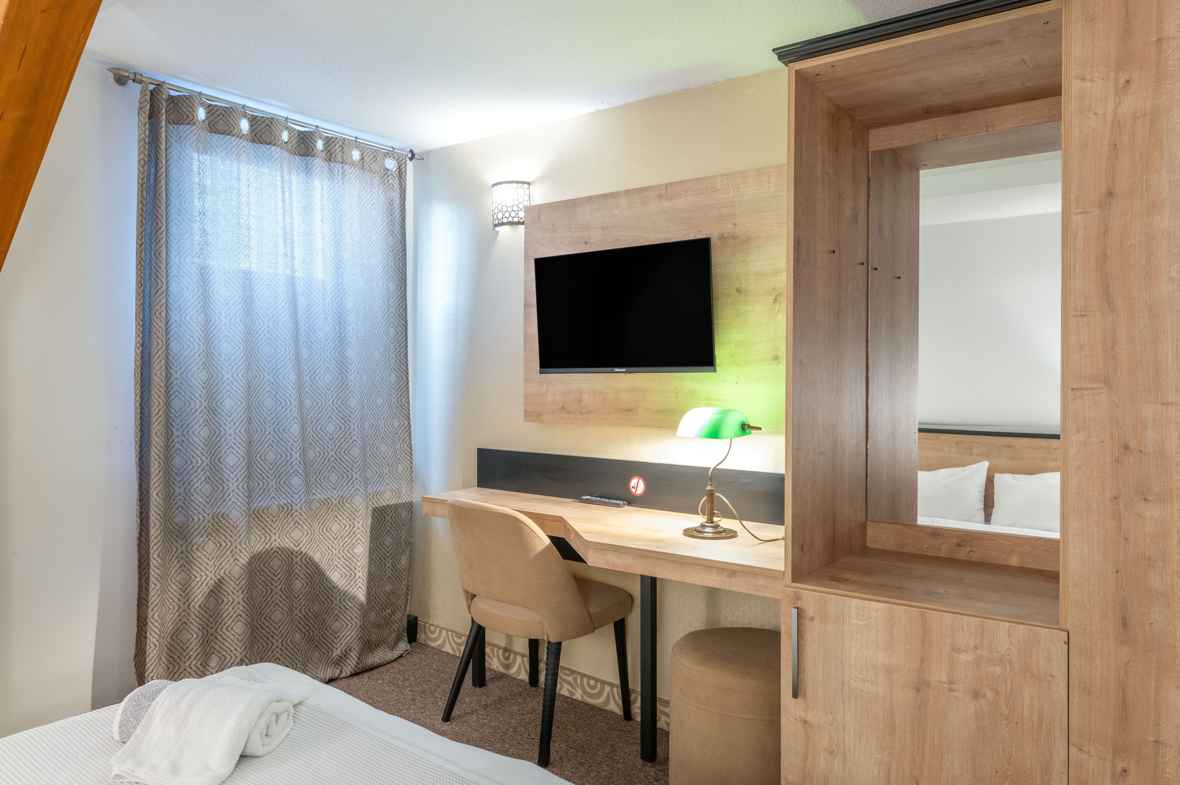 A hotel room with a bed, a desk with a chair, a television mounted on the wall, and a wardrobe.