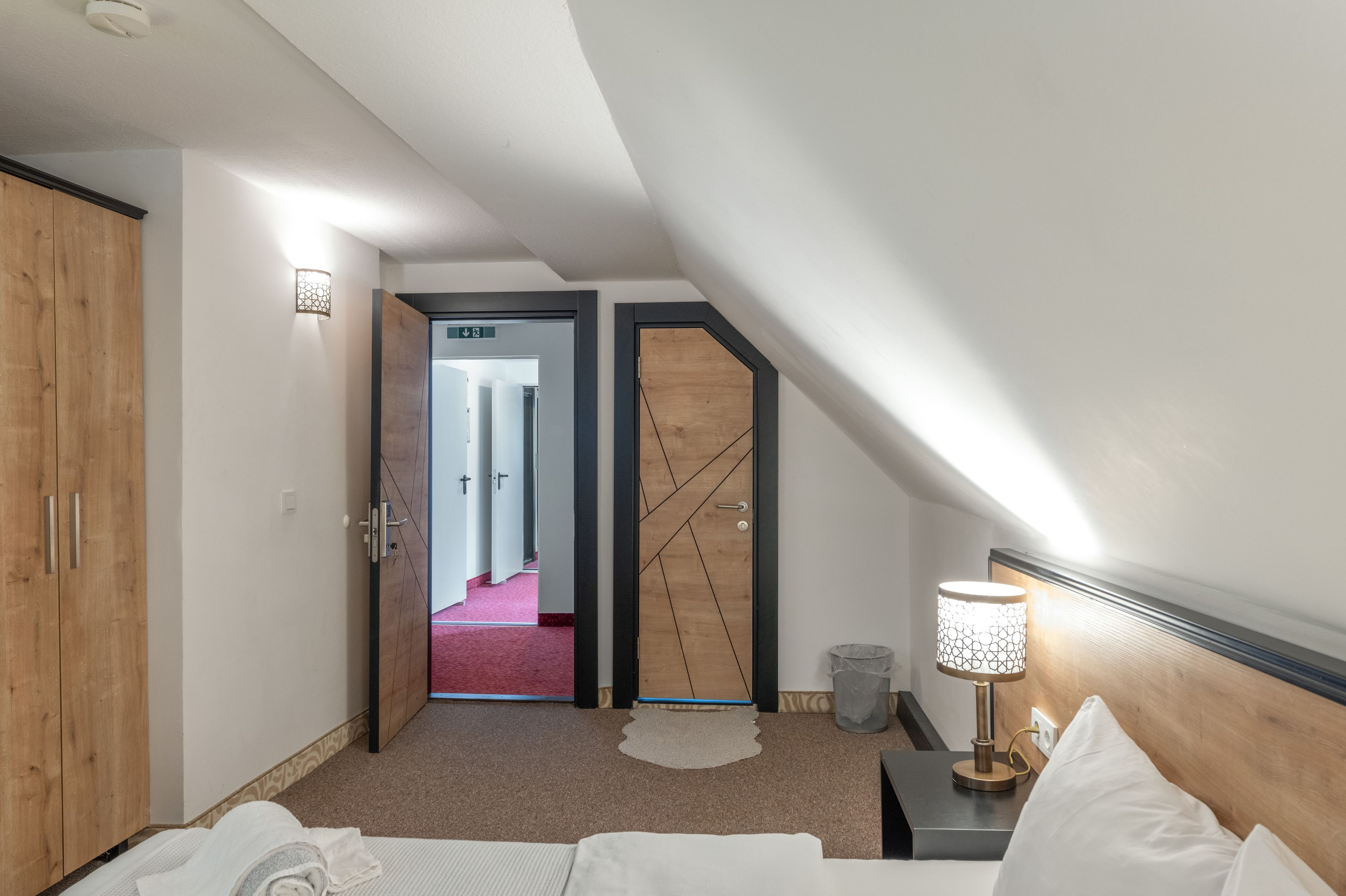Une chambre d’hôtel comprenant un lit, une table de chevet, une lampe, une armoire en bois et une porte donnant sur un couloir recouvert d’un tapis rouge.