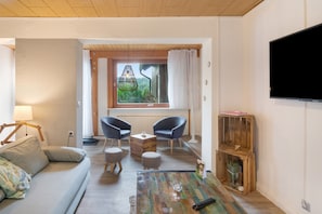 A modern living room with a sofa, two chairs, a coffee table, a TV, and a window with a view of greenery.