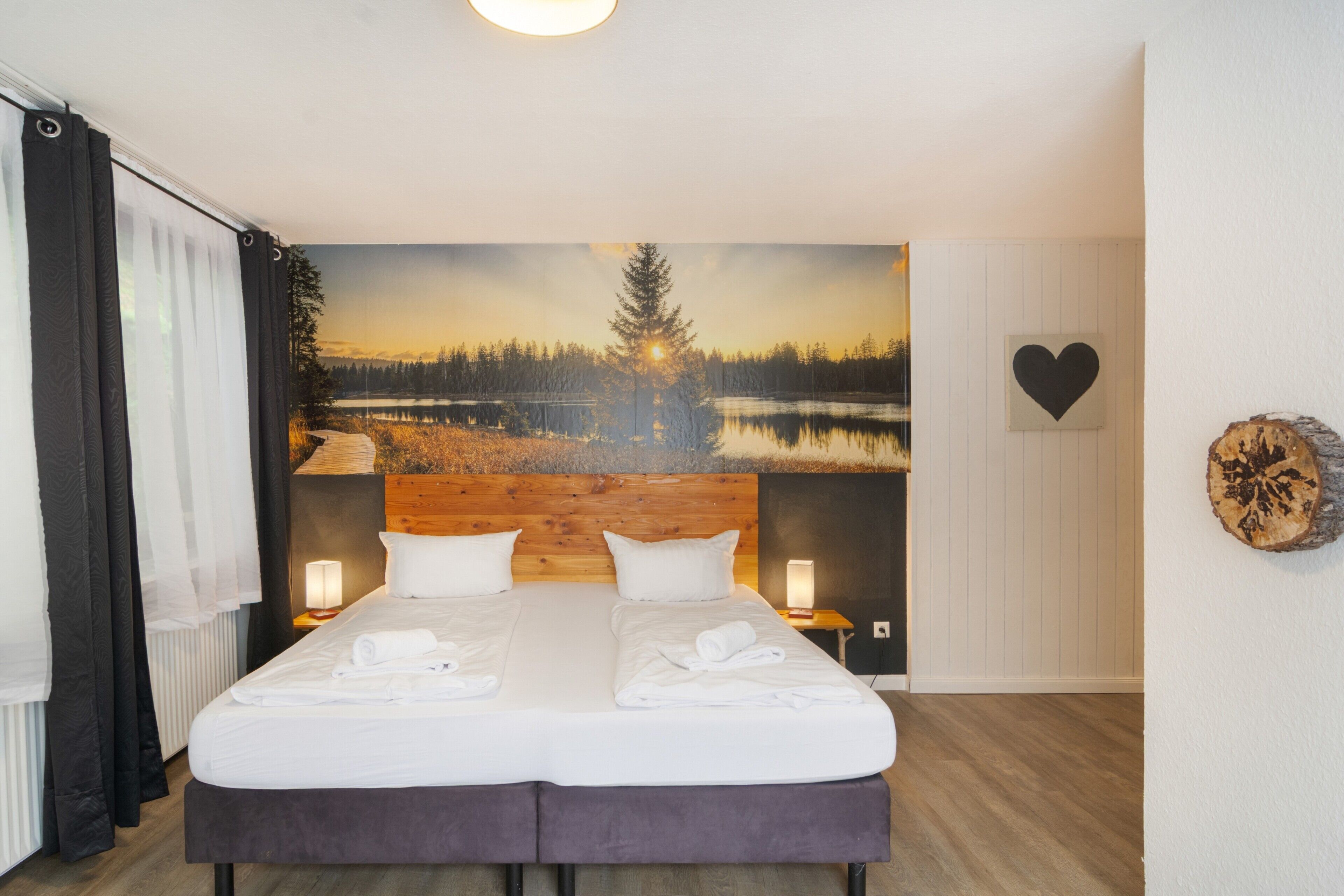 Ein Doppelbett mit weißen Leinen, zwei Nachttischlampen, ein großes Wandbild von Wald und See, eine herzförmige Dekoration und ein Holzkopfstück.