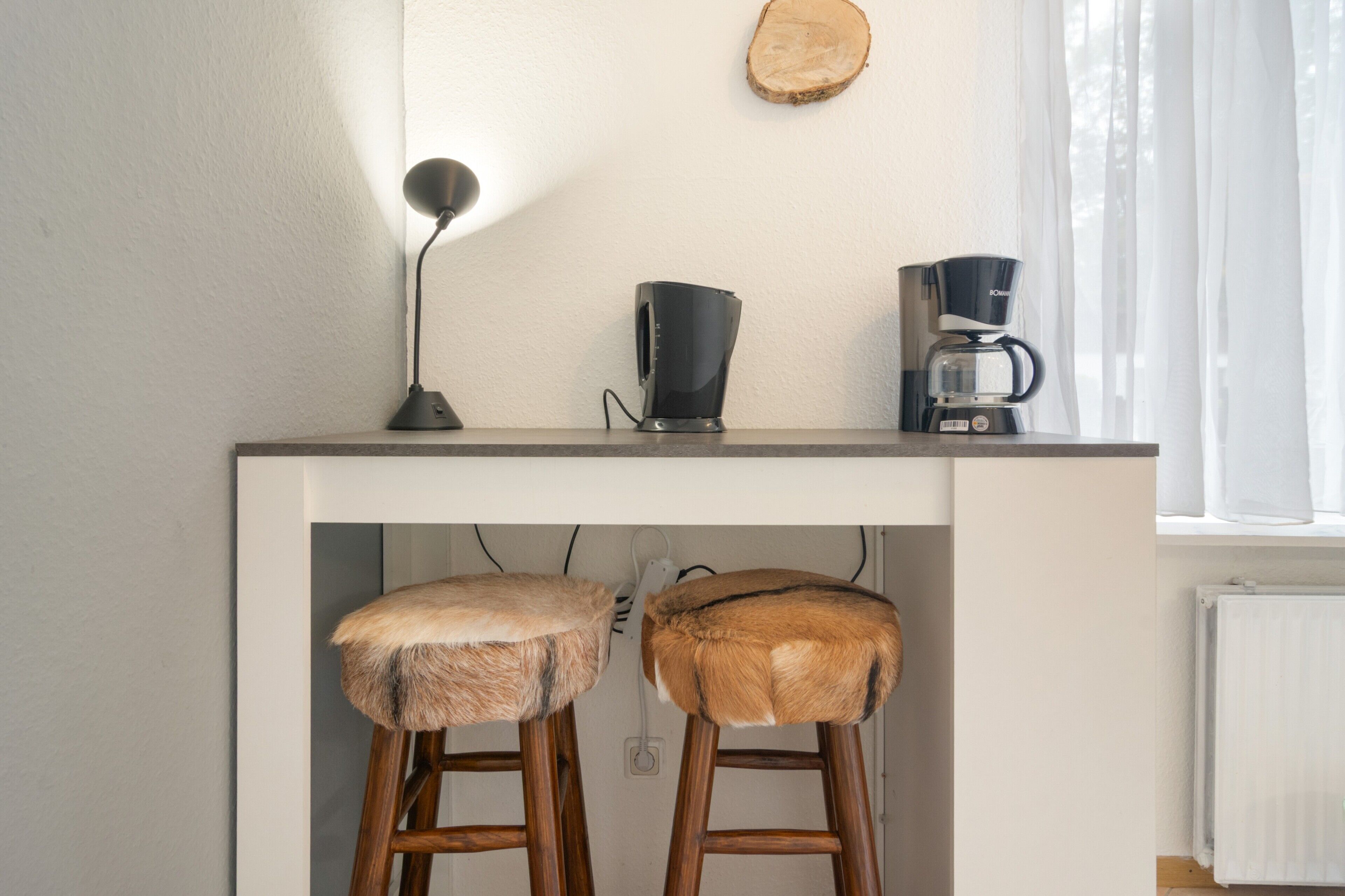 A kitchen counter with a coffee maker and a lamp, two stools with fur seats, and a wooden wall decor piece.