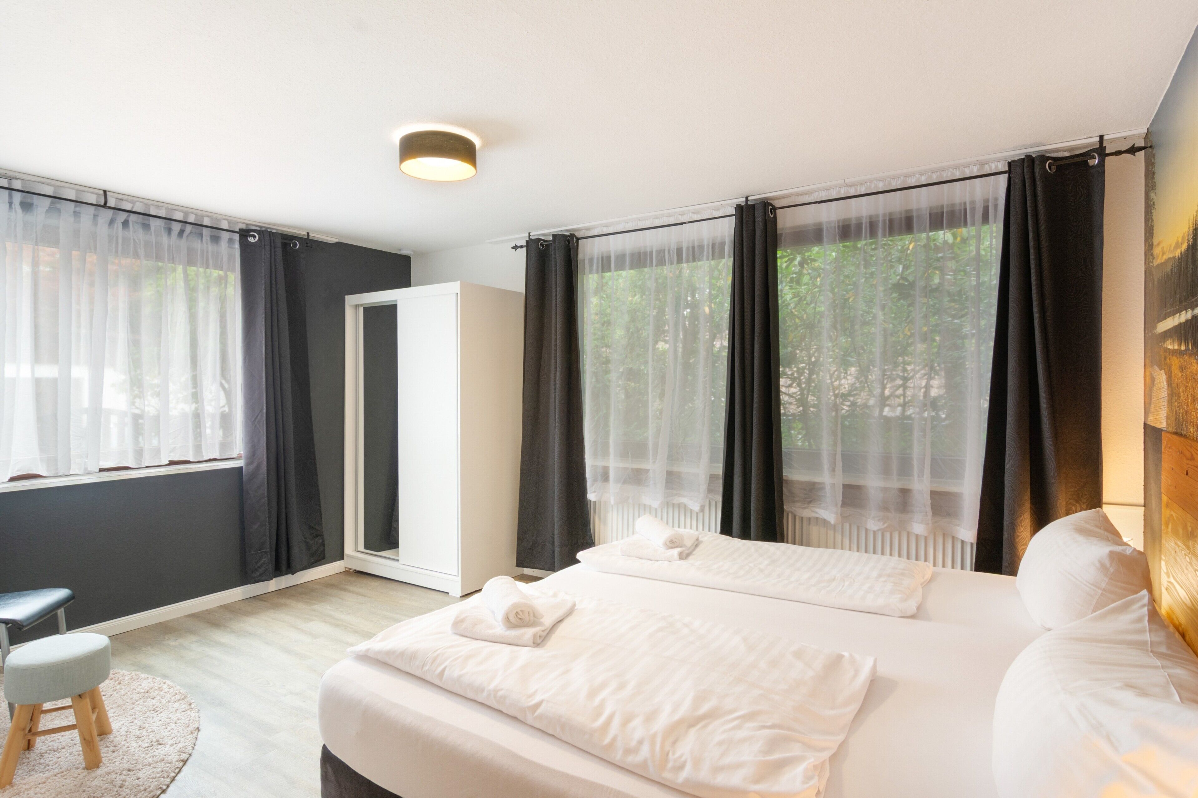 Une chambre d’hôtel avec un lit, un petit tabouret, une porte et des rideaux légers.