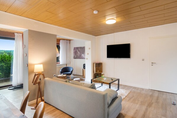 A modern living room with a sofa, a coffee table, a television mounted on the wall, and a view of the outdoors through a window.