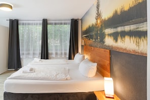 A bedroom with a large bed, a headboard, a window with curtains, and a wall-mounted lamp.