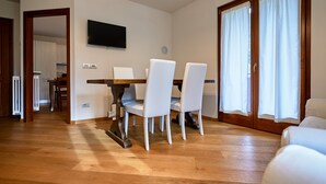 Comedor con mesa de madera y sillas blancas, televisor de pantalla plana en la pared y puerta corrediza de vidrio con cortinas.