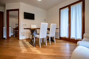 A dining area with a wooden table and white chairs, a flat-screen TV on the wall, and a sliding glass door with curtains. - Dolomis Apartments Rododendro 6 Guests (Caderzone Terme)