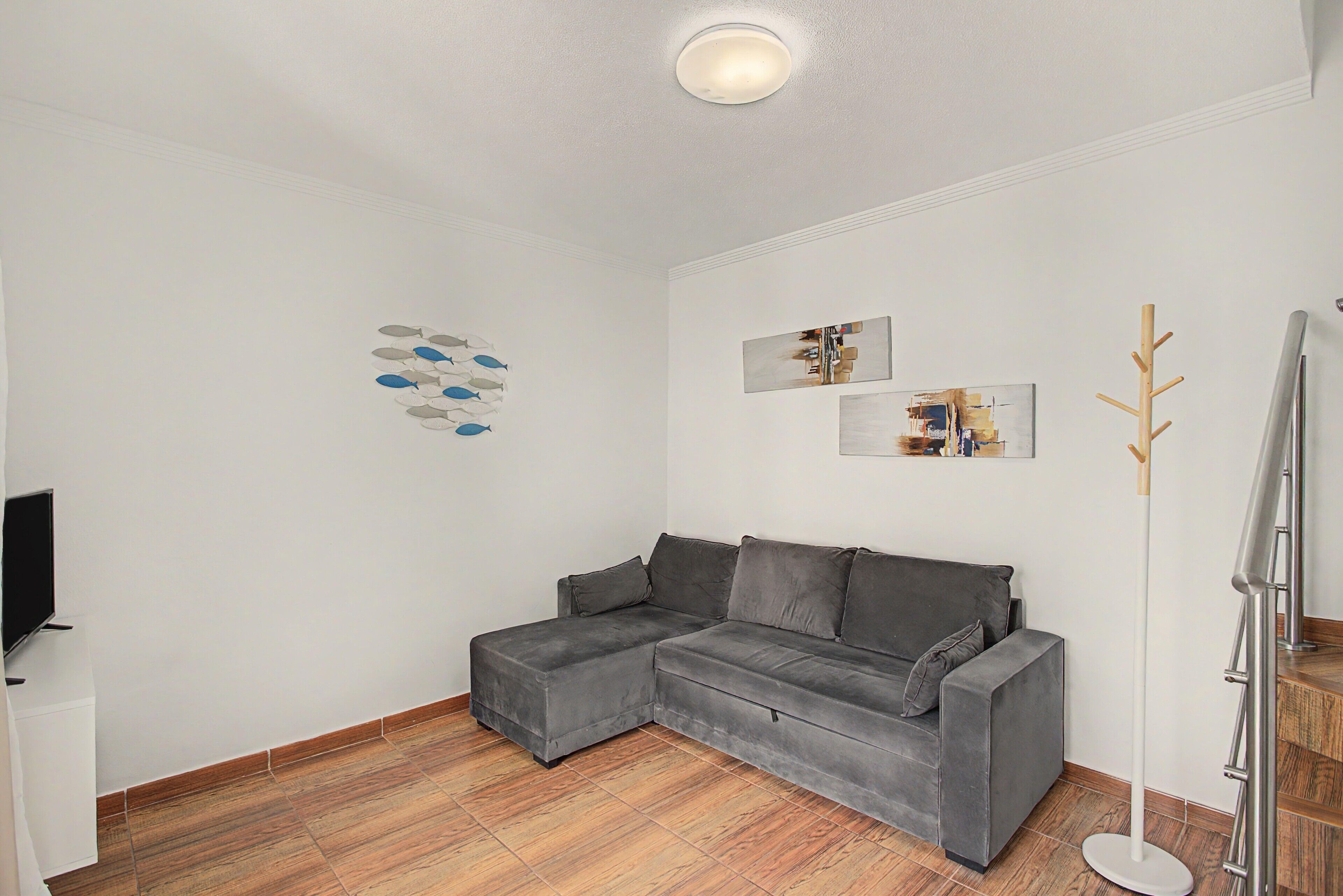 Un salon moderne avec un canapé d’angle gris, un téléviseur à écran plat, un plancher en bois et des œuvres d’art minimalistes accrochées au mur.