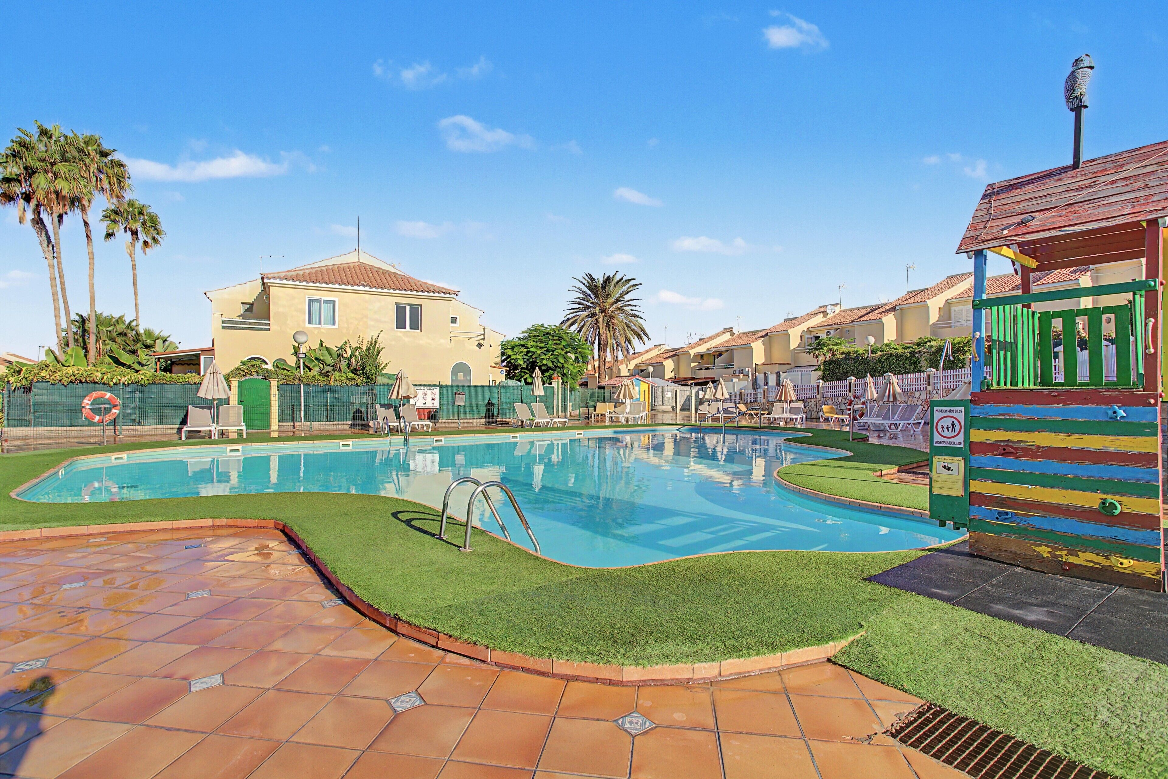 Une piscine dotée d’une terrasse carrelée, entourée de palmiers et bordée d’un bâtiment dont le toit est également recouvert de tuiles.