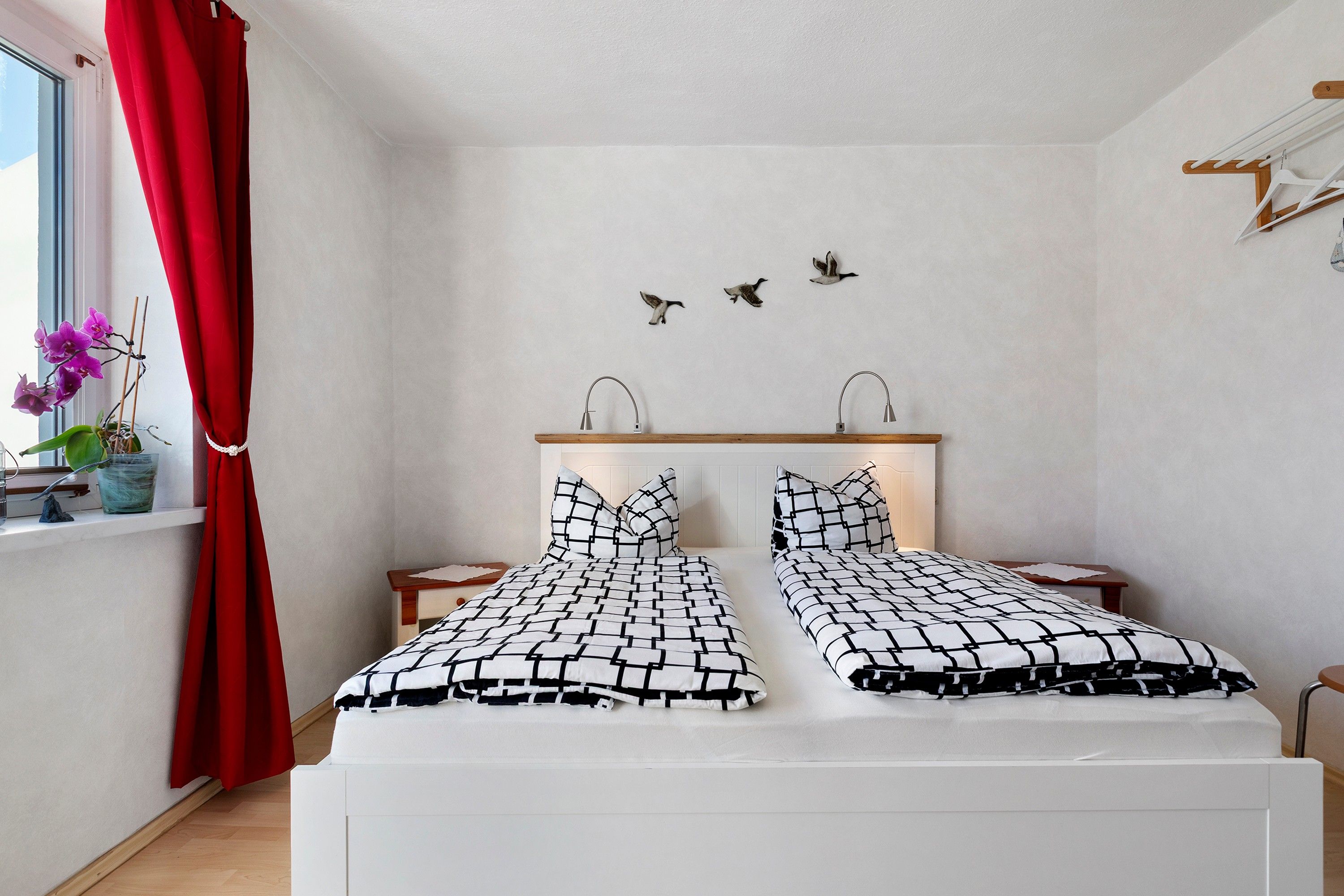 A bedroom with a bed, two nightstands, a window with a red curtain, a wall-mounted shelf, and a wall art of birds.