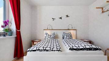 A bedroom with a bed, two nightstands, a window with a red curtain, a wall-mounted shelf, and a wall art of birds.
