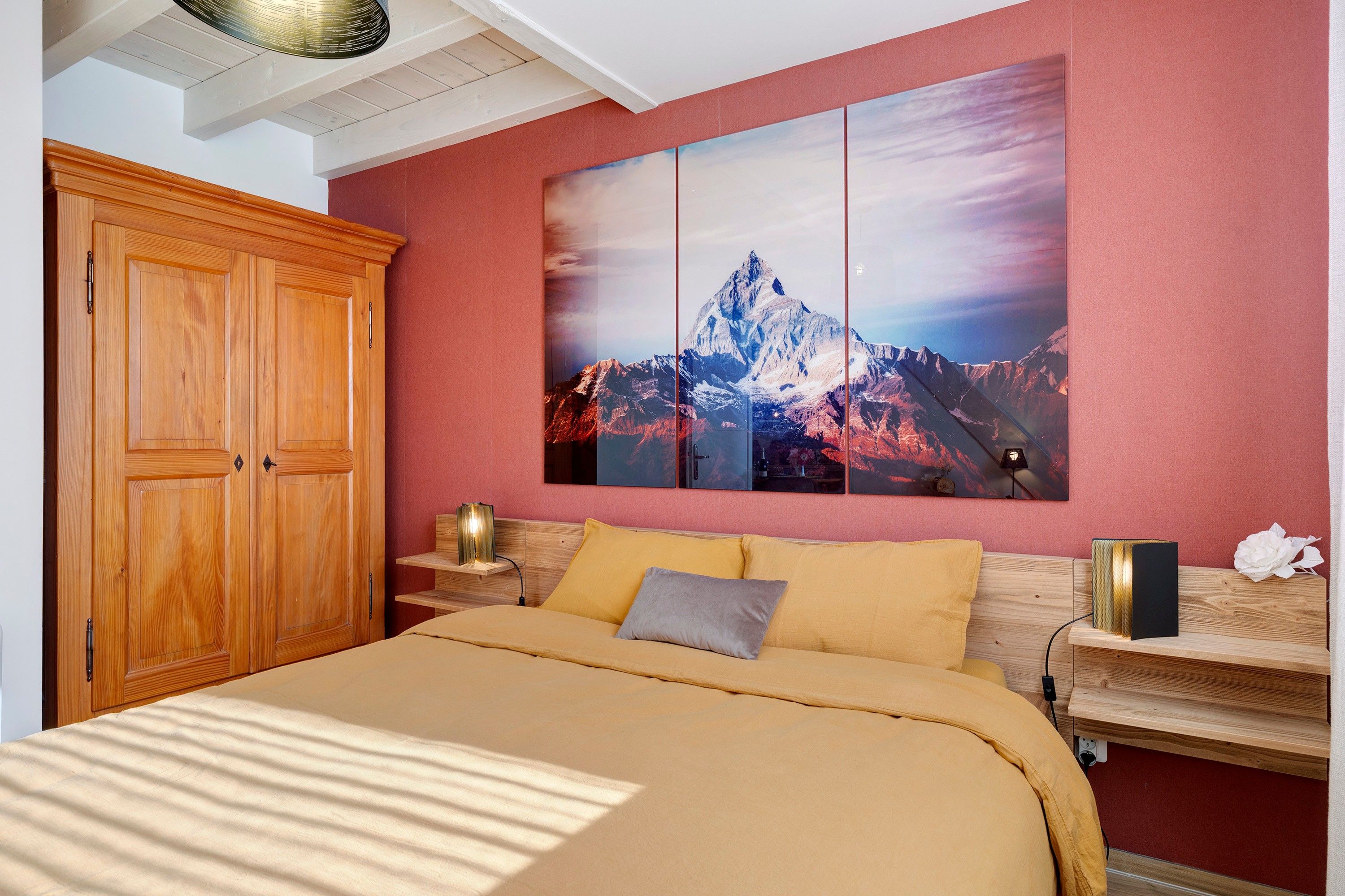 Un dormitorio con un armario de madera, una cama con cubrecama color beige, una mesita de noche con lámpara y un cuadro colgado en la pared que muestra una montaña.
