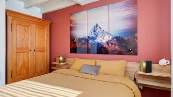 Un dormitorio con un armario de madera, una cama con cubrecama color beige, una mesita de noche con lámpara y un cuadro colgado en la pared que muestra una montaña.