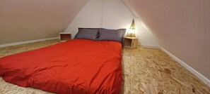 A bedroom with a sloped ceiling, a bed with a red bedspread, a nightstand with a lamp, and wooden flooring.