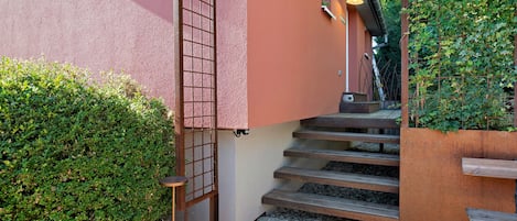 Eine Treppe mit Metallgeländer führt zu einer Tür, umgeben von Grünpflanzen und einem Kiesweg.