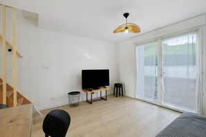 Una sala de estar moderna con un televisor de pantalla plana, un taburete pequeño, un cesto de basura y un muro de vidrio corredizo que da a un espacio exterior.