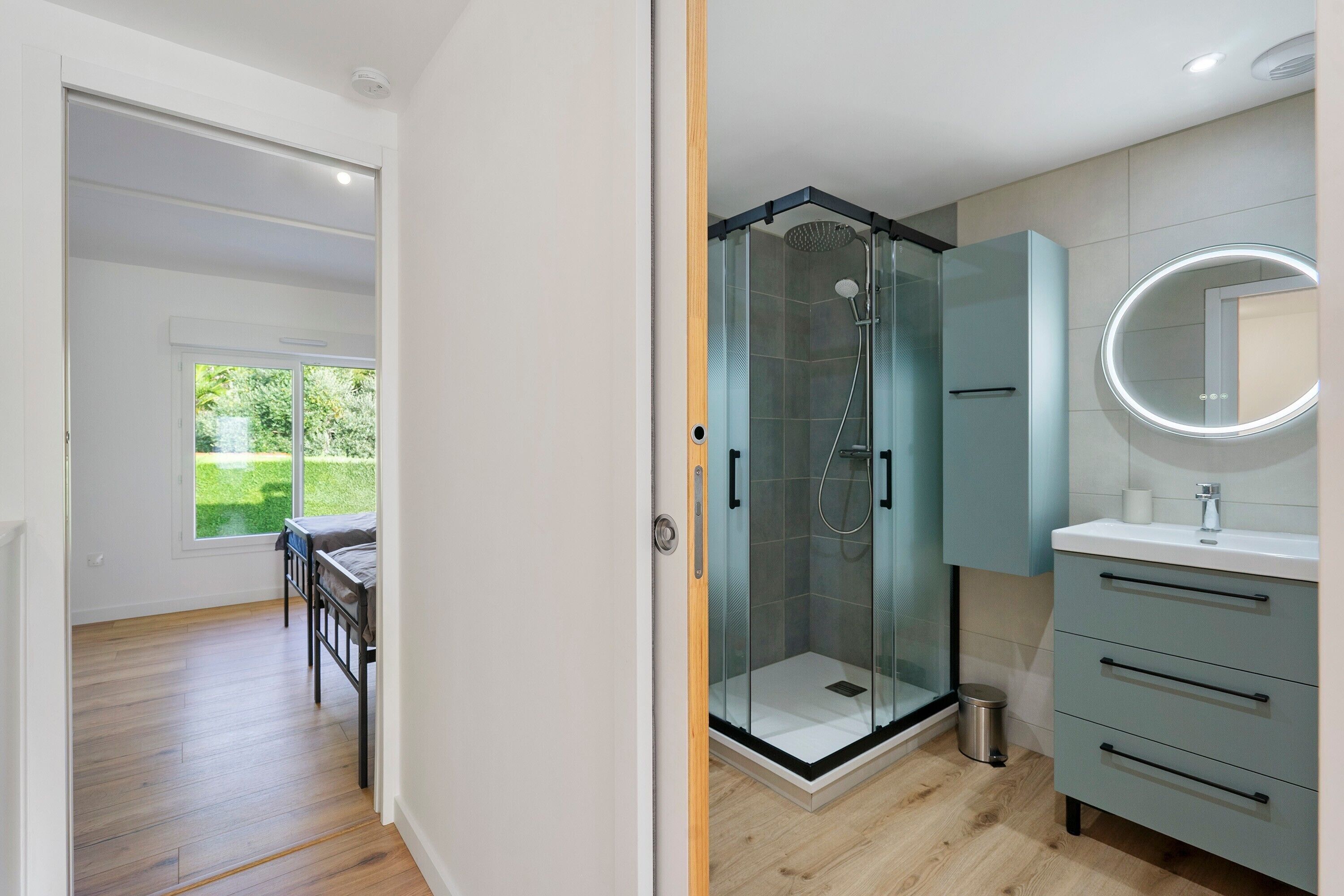 Un baño moderno con ducha envuelta en vidrio, tocador con espejo y vista a un jardín verde a través de una ventana.