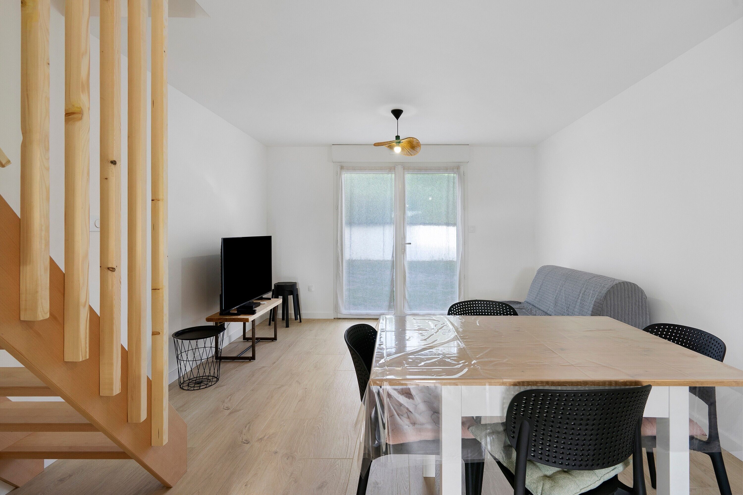 Un espacio habitable moderno y minimalista con una escalera de madera, un área de comedor con mesa y sillas, un televisor y un ventanal grande.