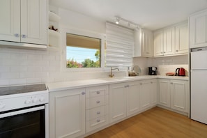 A modern kitchen with white cabinetry, a built-in oven, and a window with blinds. - Holiday Home 'Evans Garden Suite' (Nikiana)