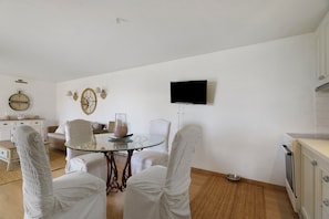 A dining area with a round glass table, white upholstered chairs, a wall-mounted television, and a clock. - Holiday Home 'Evans Garden Suite' (Nikiana)