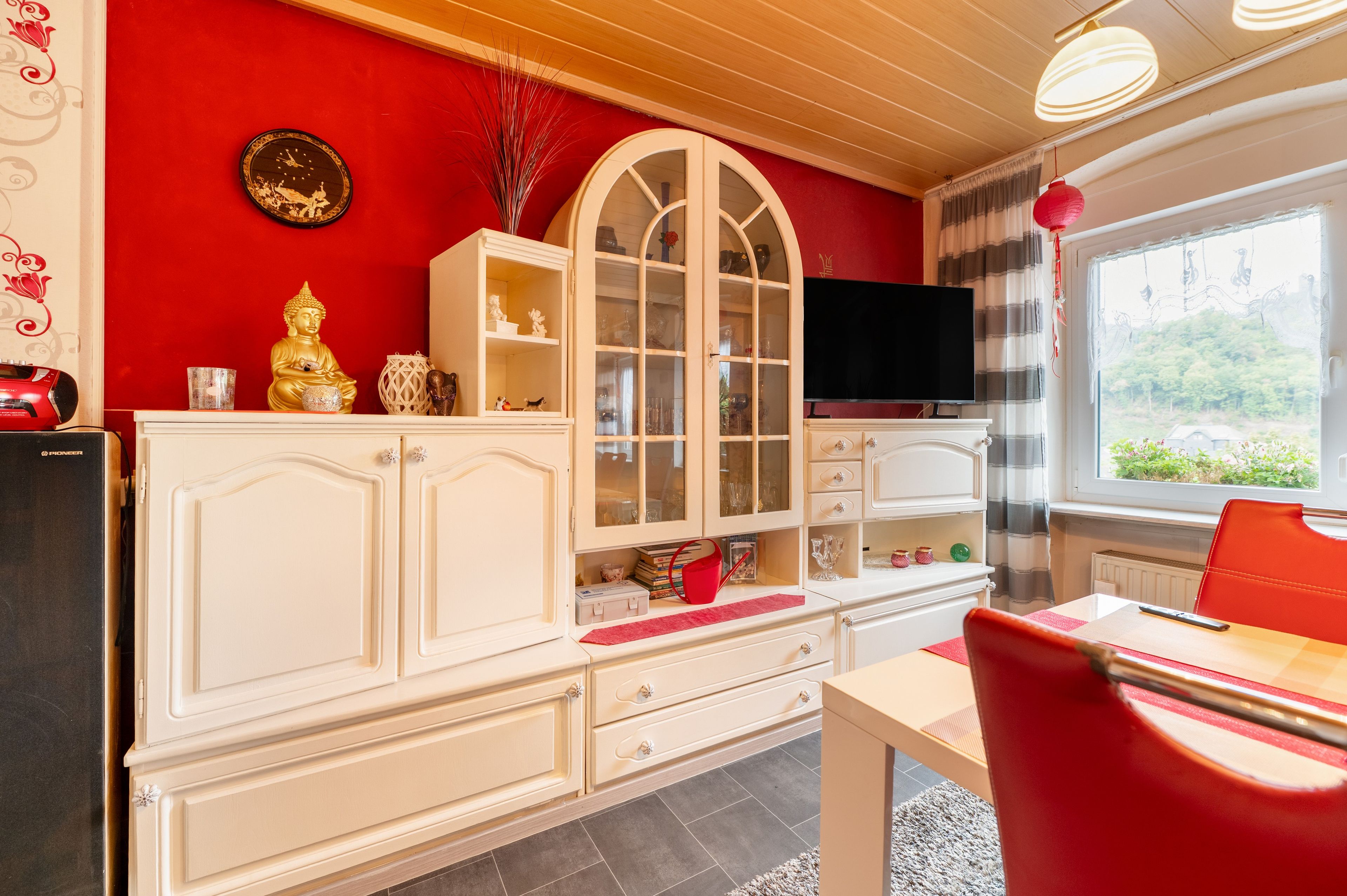 Ein Zimmer mit einer roten Wand, einem weißen Schrank mit Glastüren, einem Fernseher und einem roten Sessel.
