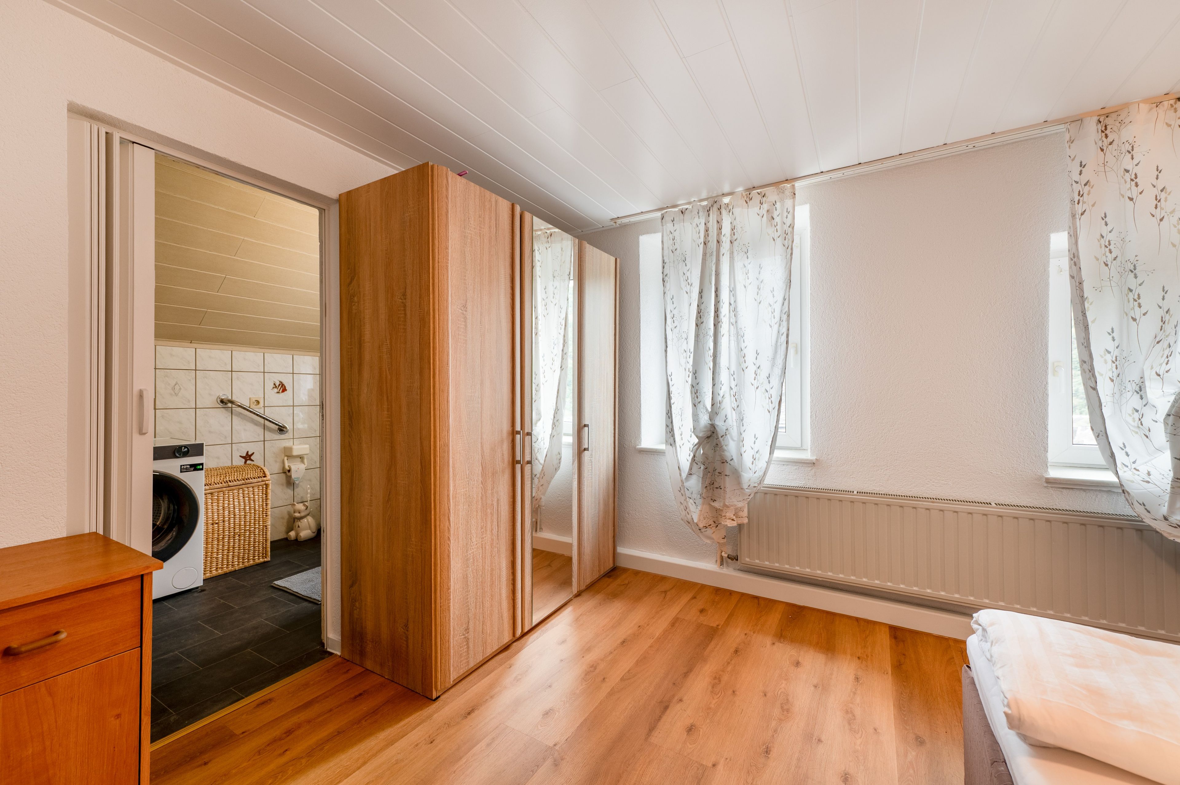 A bedroom with wooden flooring, a bed, a wardrobe, and a bathroom with a washing machine.
