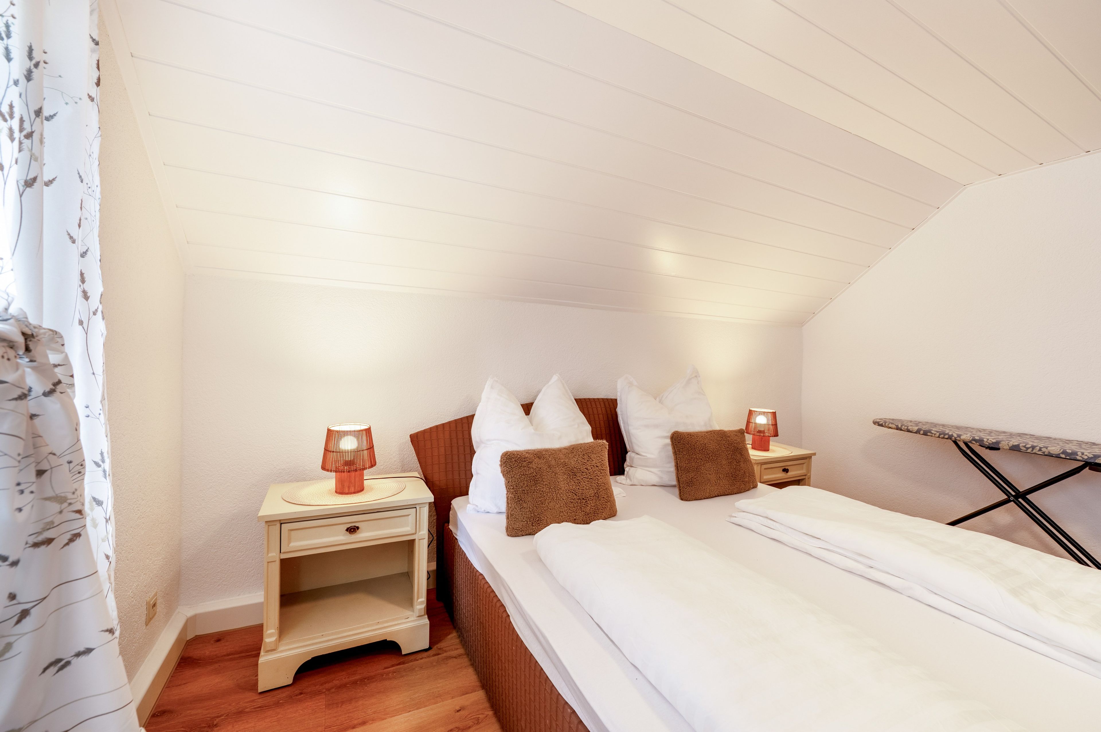 Een slaapkamer met een houten vloer, een bed met wit beddengoed, twee nachtkastjes met lampen en een raam met gordijnen.