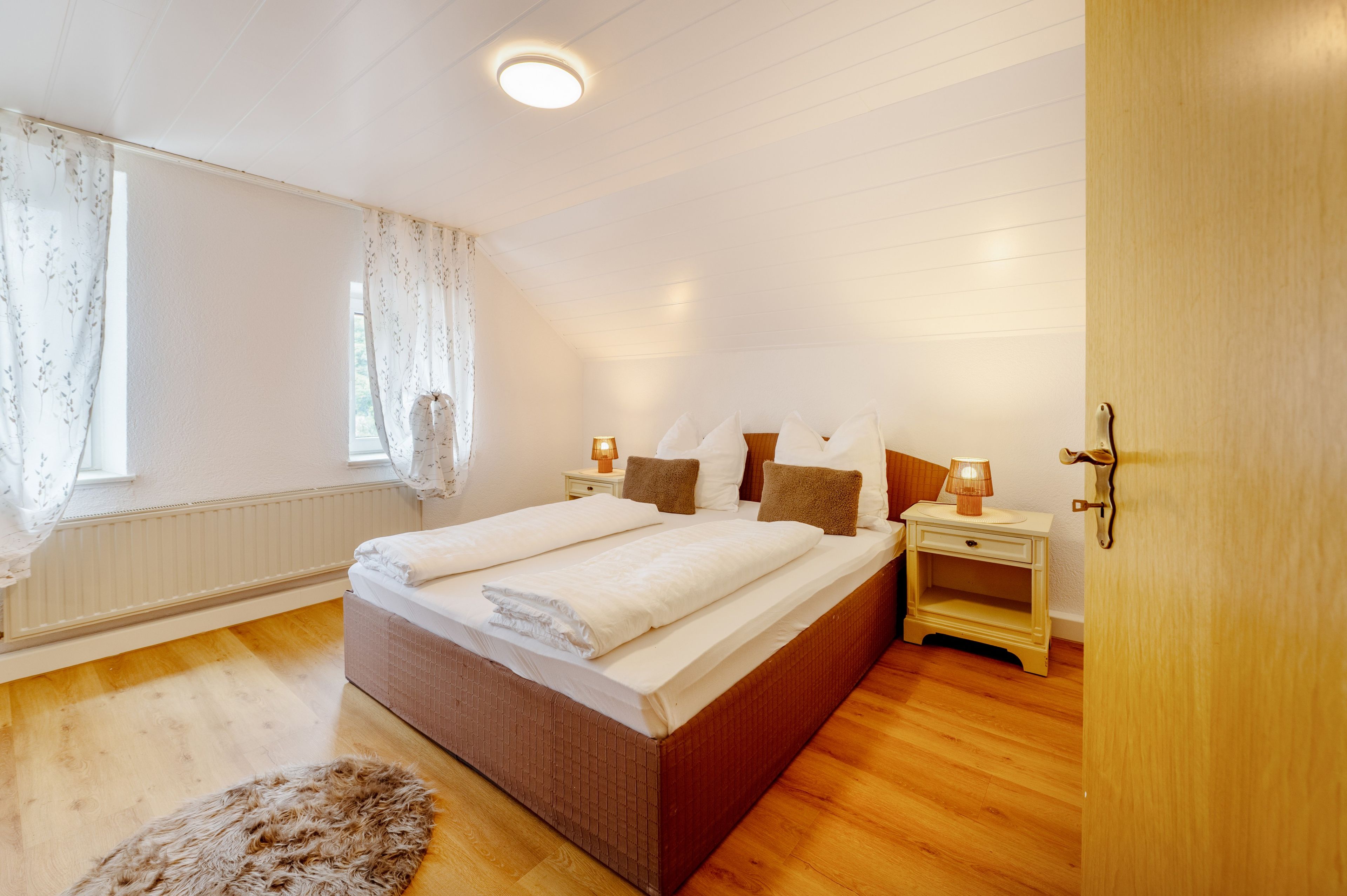 A bedroom with a large bed, two bedside tables with lamps, a wooden door, and a window with sheer curtains.
