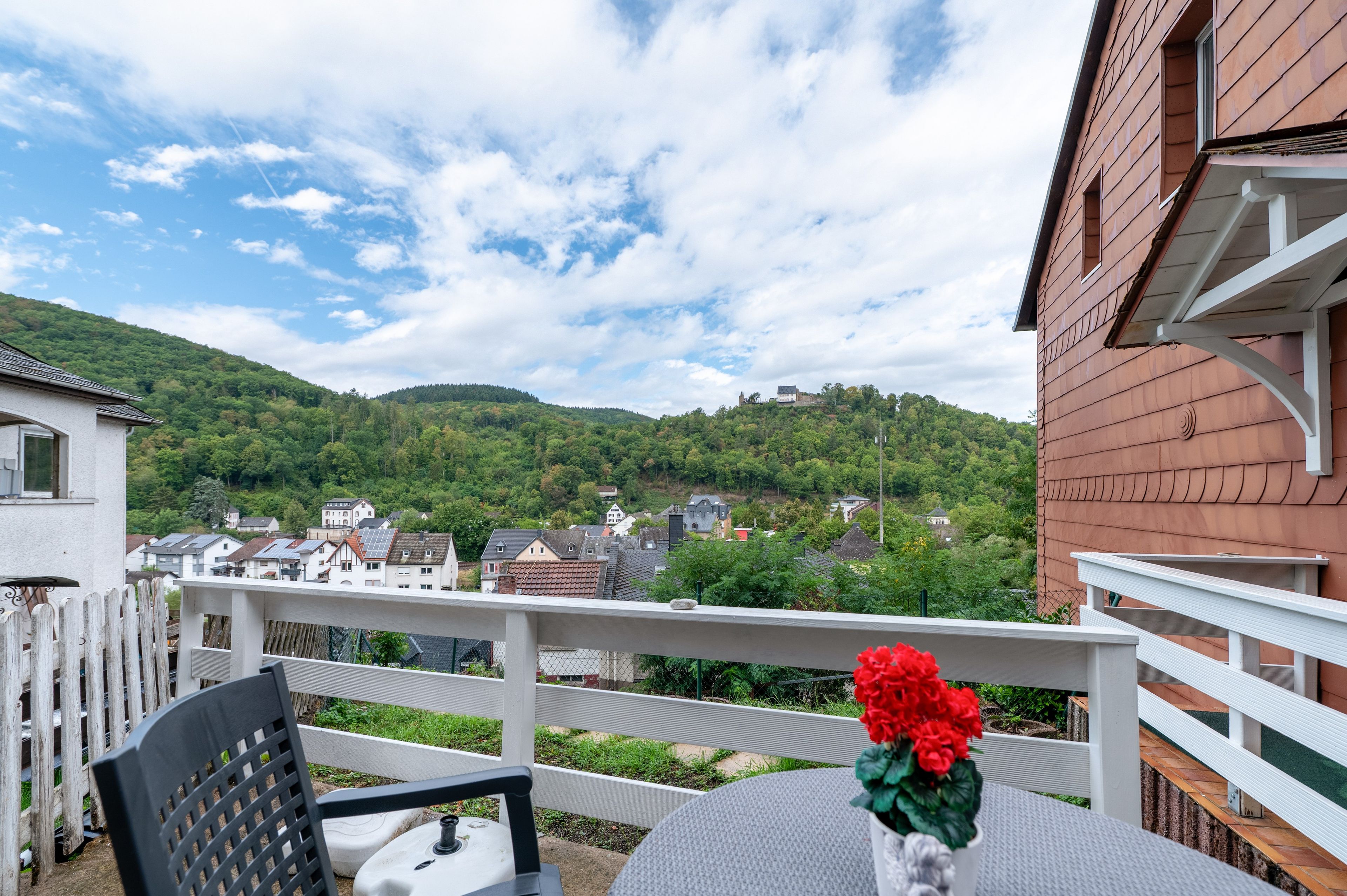 A balcony with a view of a hillside village, a white railing, and a table with a chair.